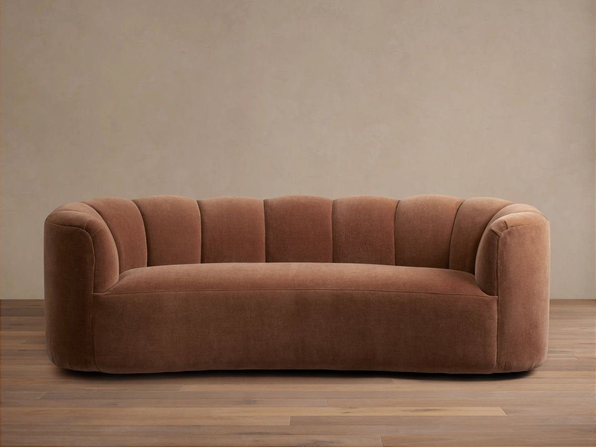 Warm brown Amira Sofa with fluted, curved back and integrated arms, on a wooden floor against a neutral wall.