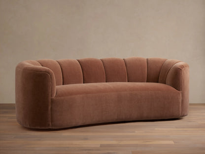 Curved Amira Sofa in muted terracotta velvet, featuring a channel-tufted back and rounded arms, on a wood floor.