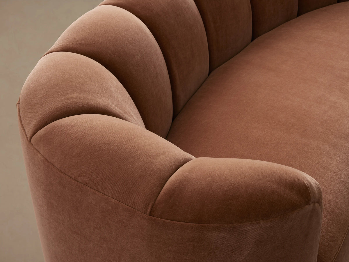 Amira Sofa, back view. Curved, reddish-brown velvet sofa with tufted detailing along the top of the backrest against a light wall.