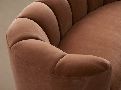 Amira Sofa, back view. Curved, reddish-brown velvet sofa with tufted detailing along the top of the backrest against a light wall.