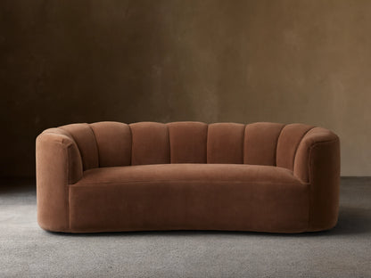 Warm brown Amira Sofa with fluted, curved back and integrated arms, on a wooden floor against a neutral wall.