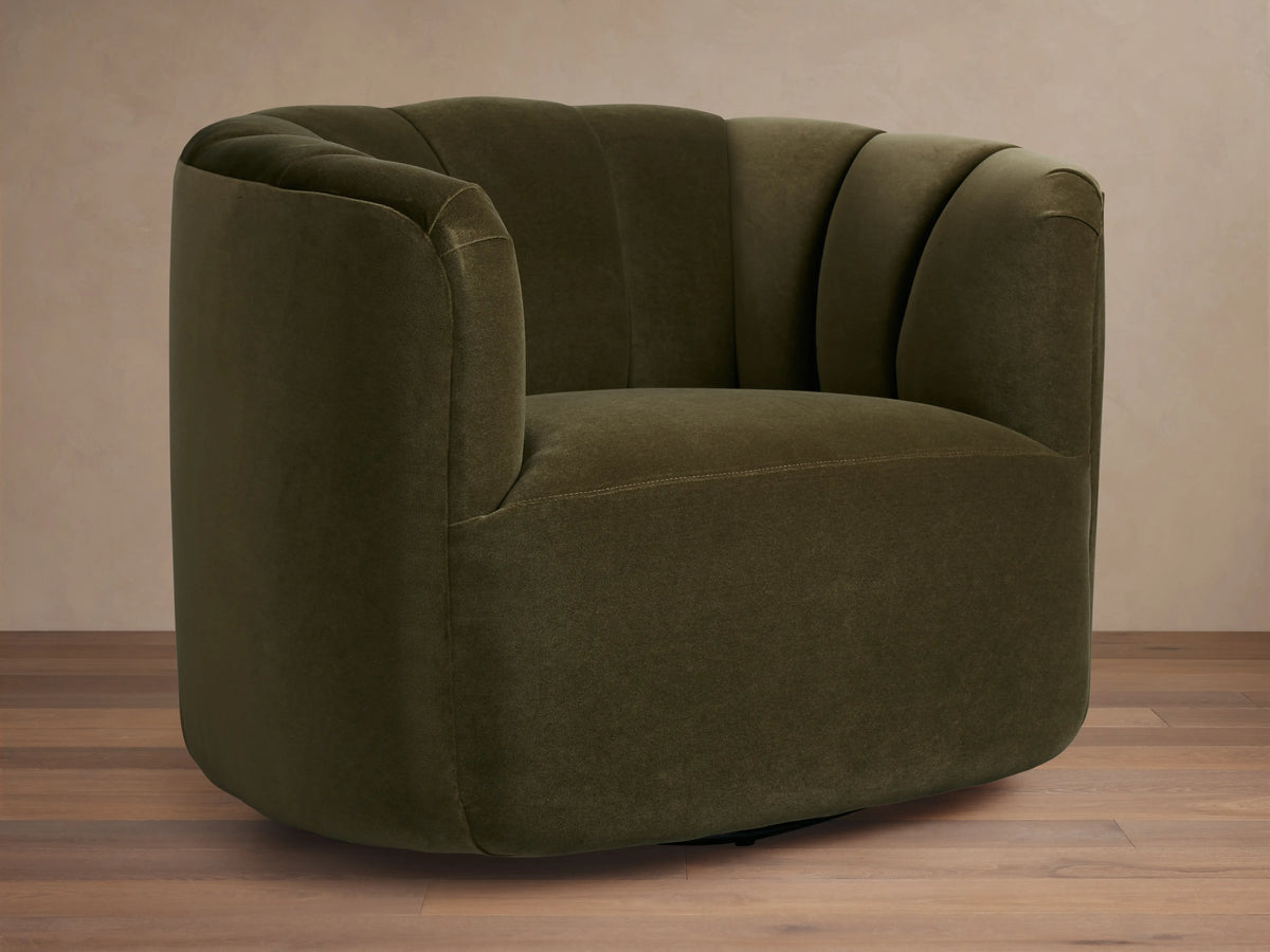 Dark olive green velvet Amira Swivel Chair, rounded form, vertical channel tufting on back, angled view on wood floor.