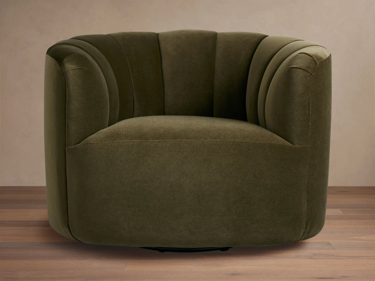 Dark olive green velvet Amira Swivel Chair, rounded form, vertical channel tufting on back, angled view on wood floor.