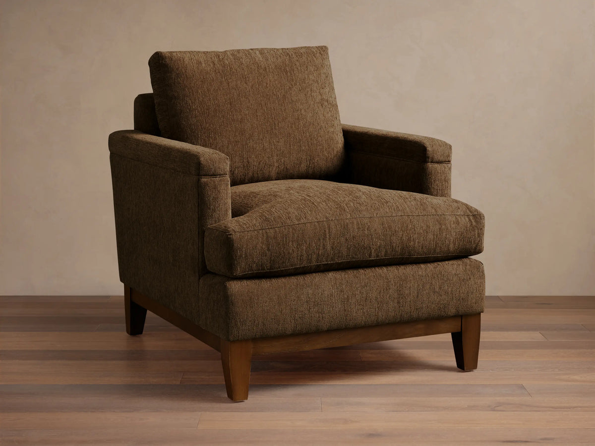 Front view of a brown textured fabric armchair with wide arms, loose cushions, and a visible dark wood base and legs.