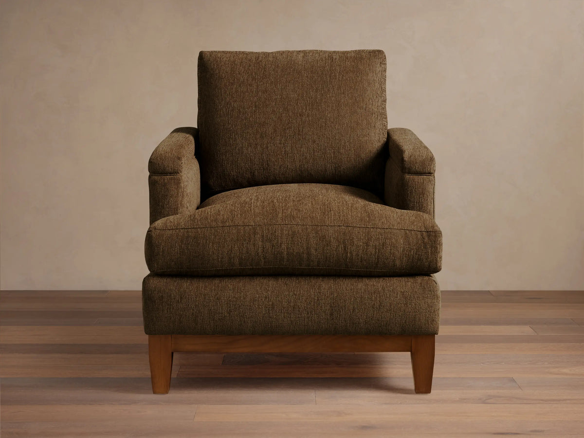 Front view of a brown textured fabric armchair with wide arms, loose cushions, and a visible dark wood base and legs.