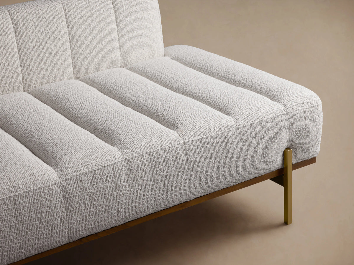 Lansing Daybed with soft beige upholstery, tufted design, and minimalist brass legs on a wooden floor, against a beige wall.