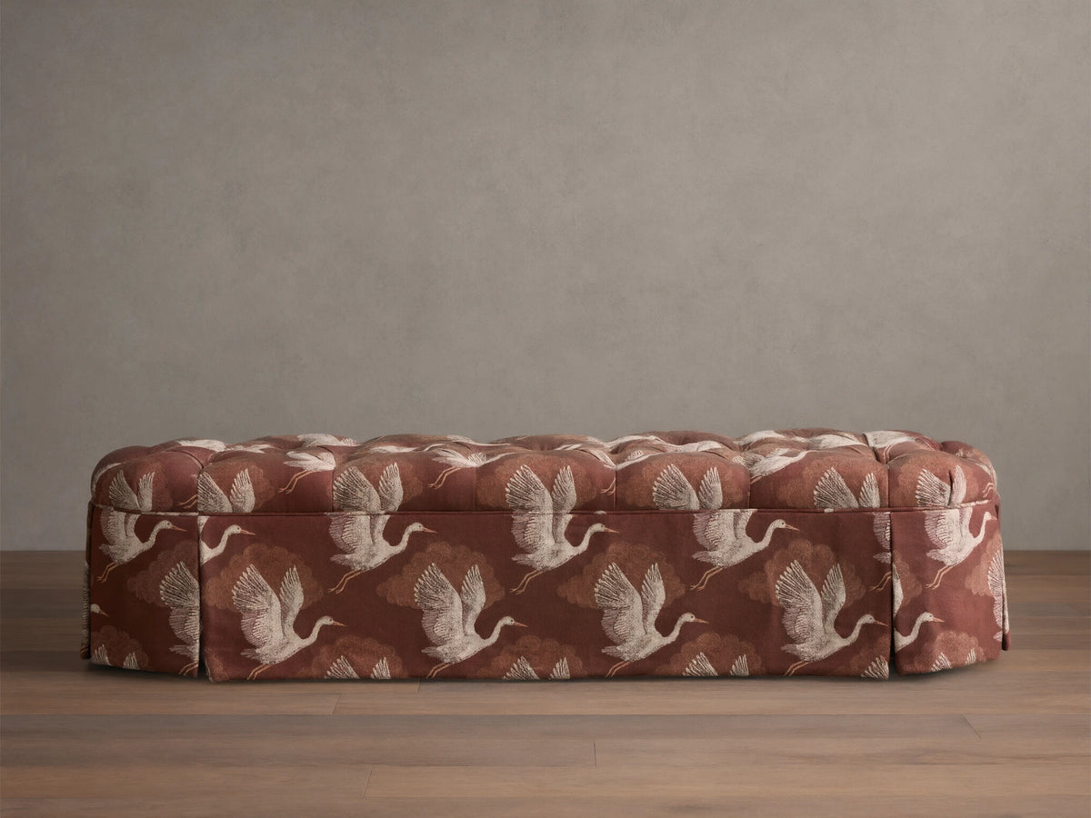 Front view of the Liddie Oval Bench with red-brown fabric, a tufted top, and a white flying crane pattern with a pleated skirt.