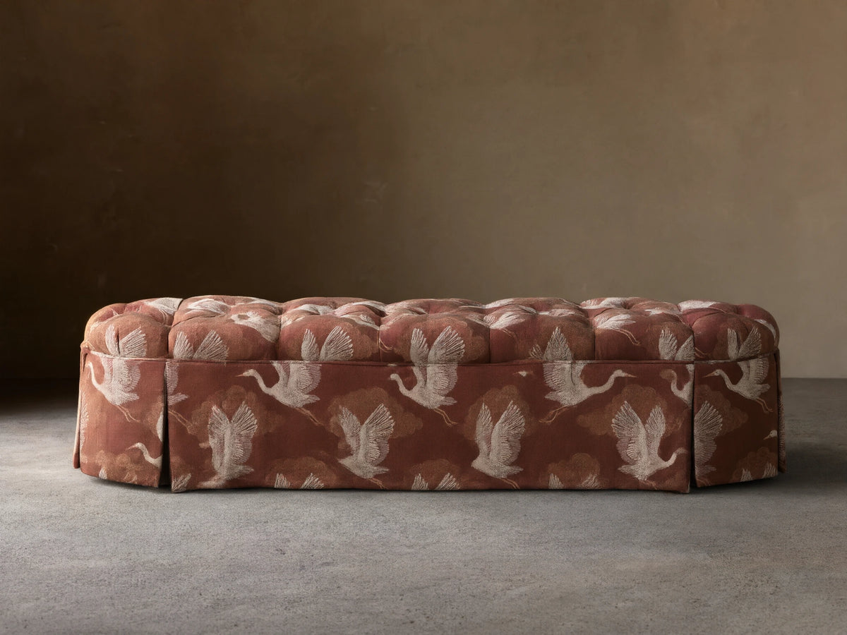 Front view of the Liddie Oval Bench with red-brown fabric, a tufted top, and a white flying crane pattern with a pleated skirt.