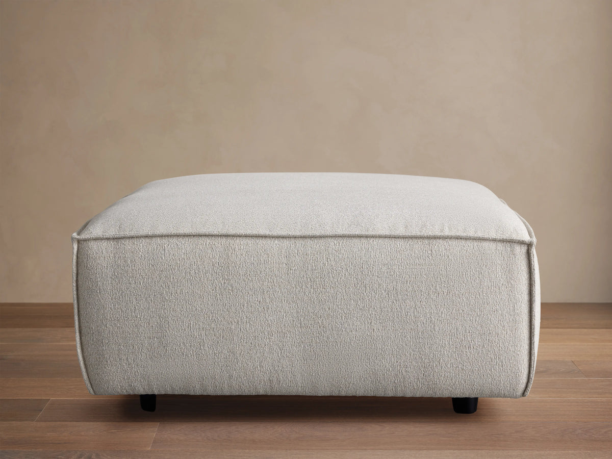 Front view of the light grey Coburn Ottoman, featuring a textured fabric and precise piping details on its rounded edges.