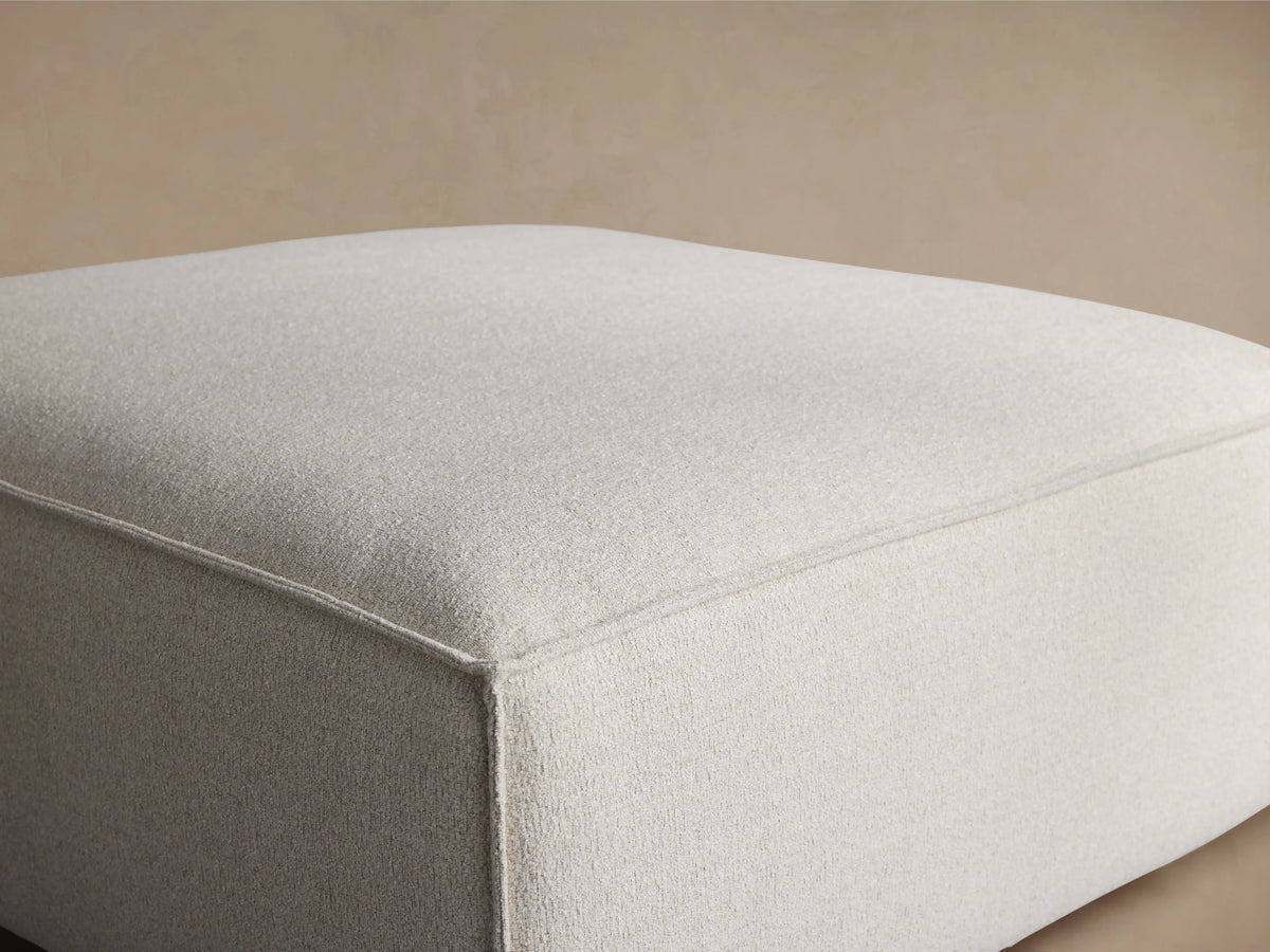 Angled view of a light beige Coburn Ottoman, featuring textured fabric and tailored piping along the seams.