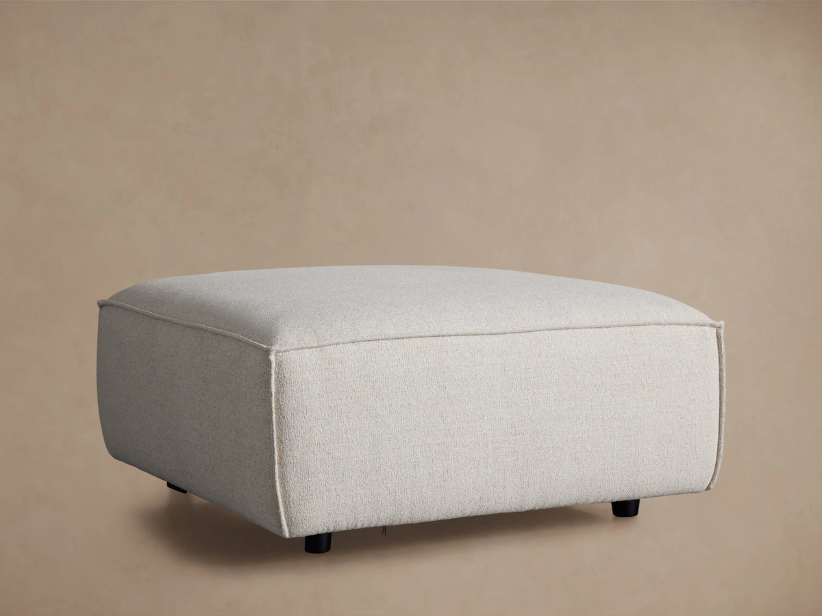 Front view of the light grey Coburn Ottoman, featuring a textured fabric and precise piping details on its rounded edges.