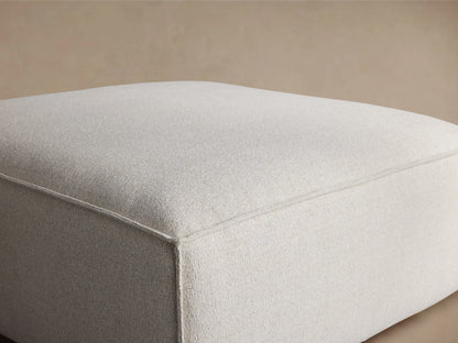 Front view of the oatmeal-colored upholstered Coburn Ottoman, featuring clean lines, visible piping, and small dark legs.