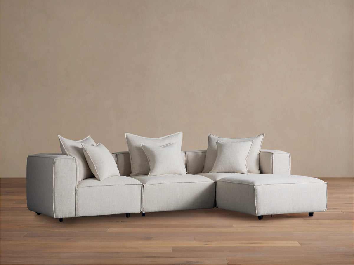 Alt text: Coburn Four-Piece Sectional in neutral fabric on a wooden floor, featuring three seating sections and an attached chaise with plush cushions.