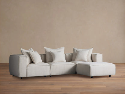 Alt text: Coburn Four-Piece Sectional in neutral fabric on a wooden floor, featuring three seating sections and an attached chaise with plush cushions.