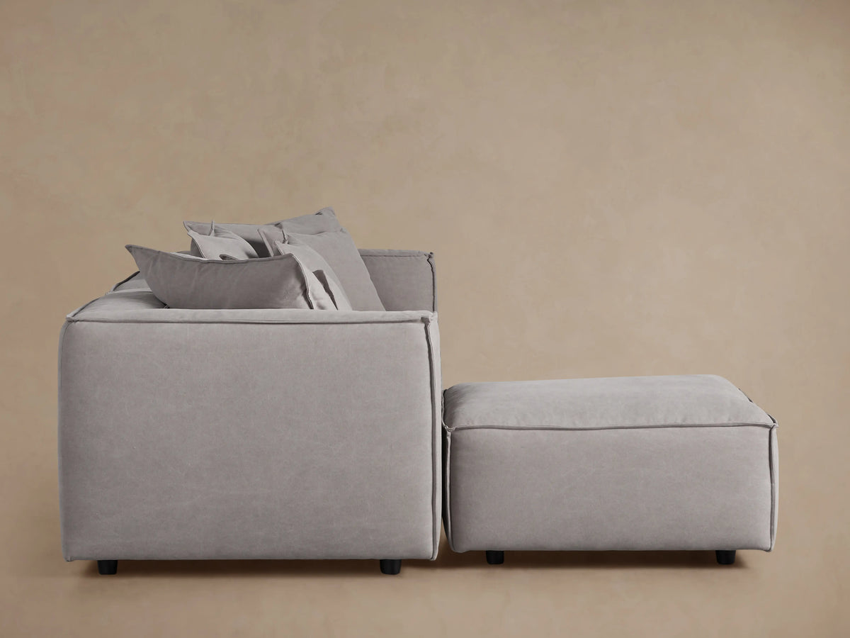 Light gray Coburn Four-Piece Sectional sofa with plush cushions and matching ottoman, viewed from the side against a beige wall.
