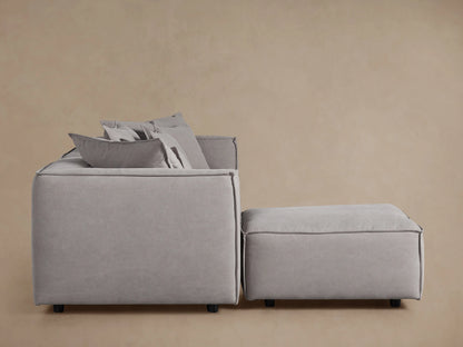 Light gray Coburn Four-Piece Sectional sofa with plush cushions and matching ottoman, viewed from the side against a beige wall.
