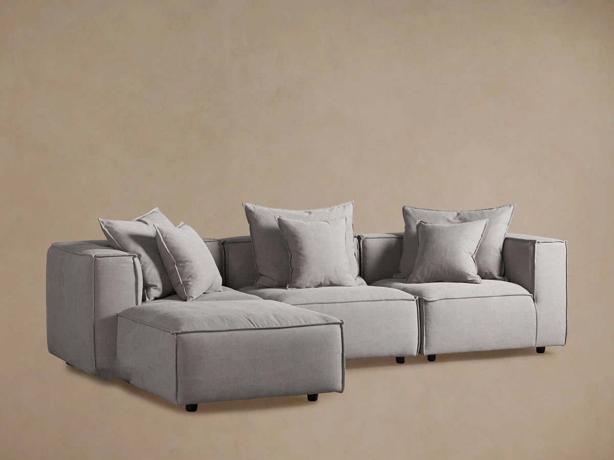 Light grey Coburn Four-Piece Sectional sofa, modular design with boxy cushions & accent pillows, on wood floor, beige wall.