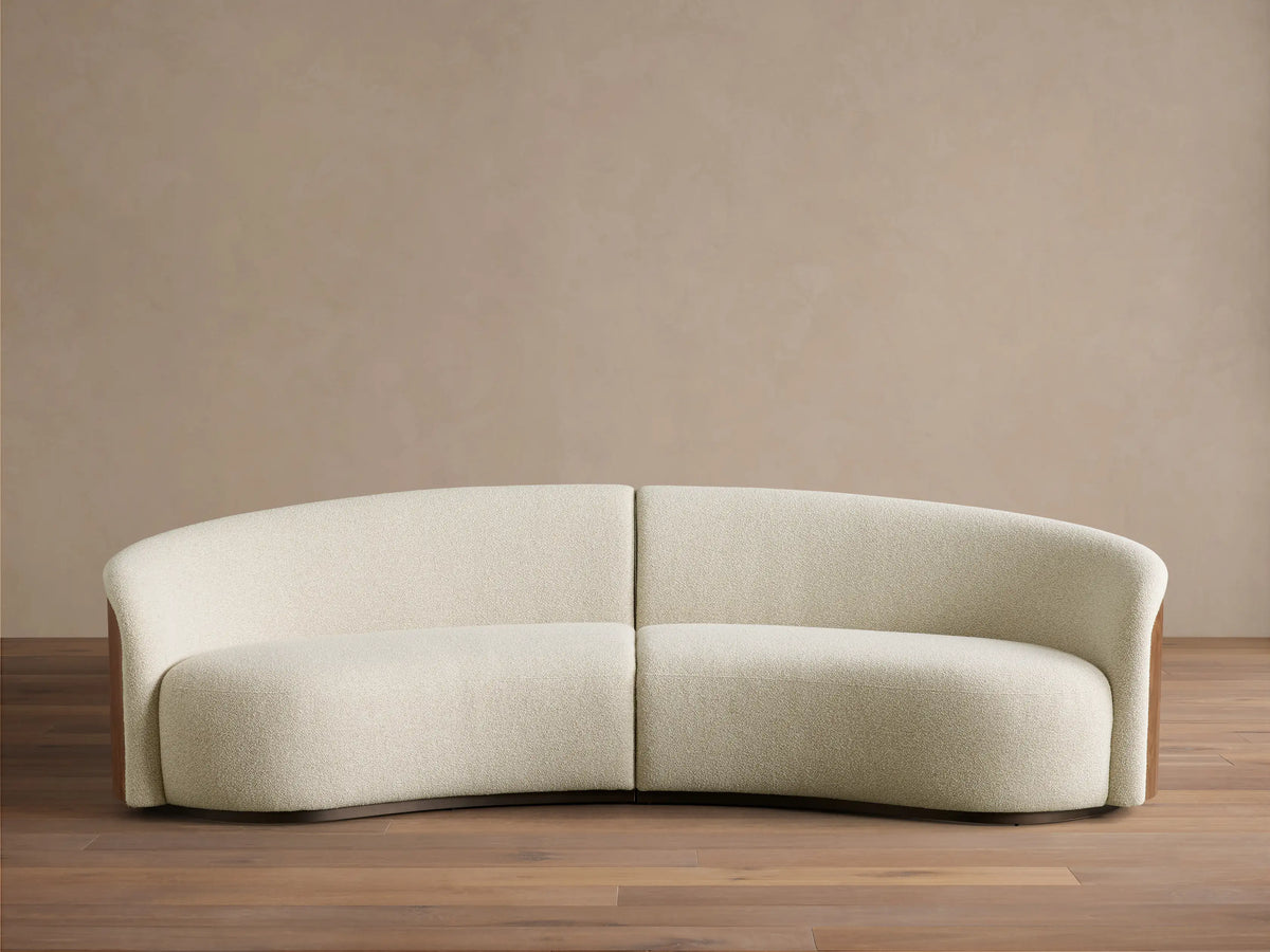 Front view of the curved Caden Two-Piece Sectional, upholstered in cream textured fabric with dark walnut wood veneer sides.