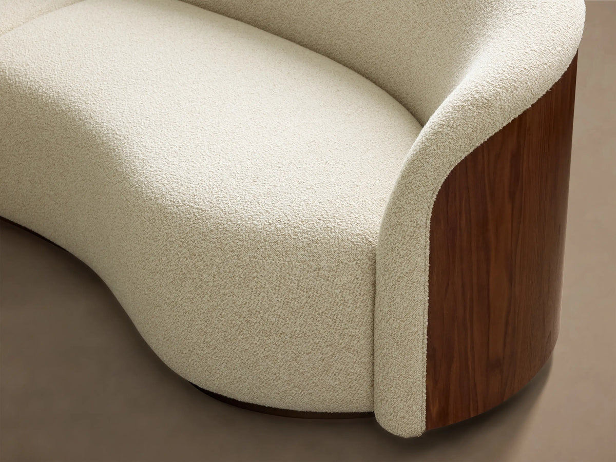 Close-up of the curved light beige textured upholstery and dark walnut wood panel on the Caden Three-Piece Bumper Sectional.