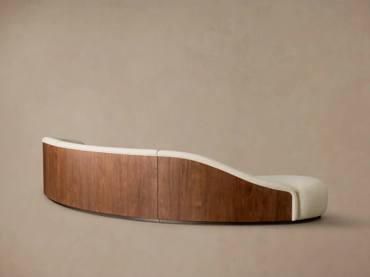 Rear and side view of a curved Caden Three-Piece Bumper Sectional, featuring cream upholstery and walnut wood veneer.