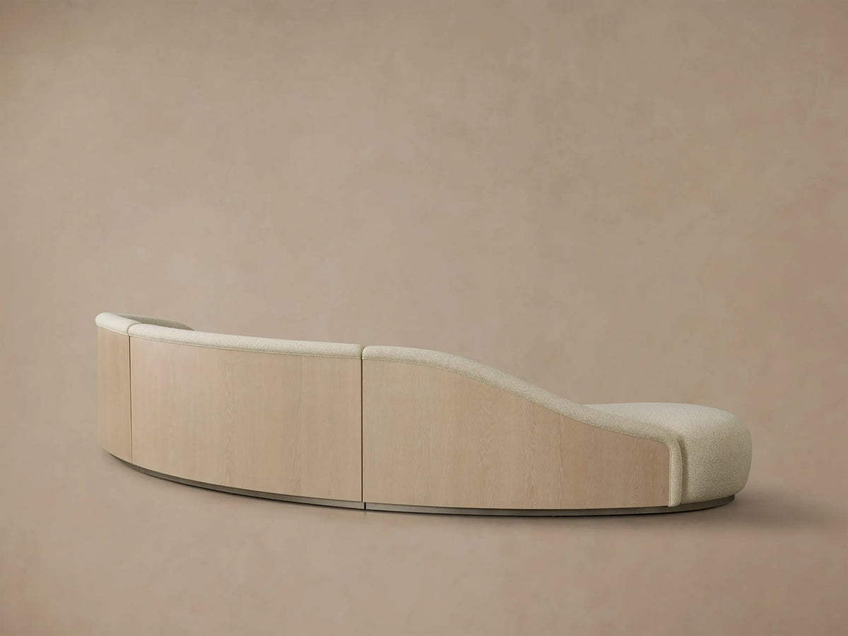 Rear view of the curved Caden Three-Piece Bumper Sectional with light sand-colored fabric, oak veneer, and a brushed nickel plinth.