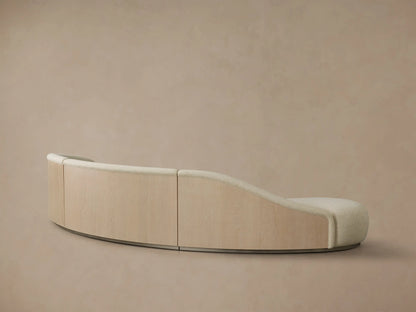 Rear view of the curved Caden Three-Piece Bumper Sectional with light sand-colored fabric, oak veneer, and a brushed nickel plinth.