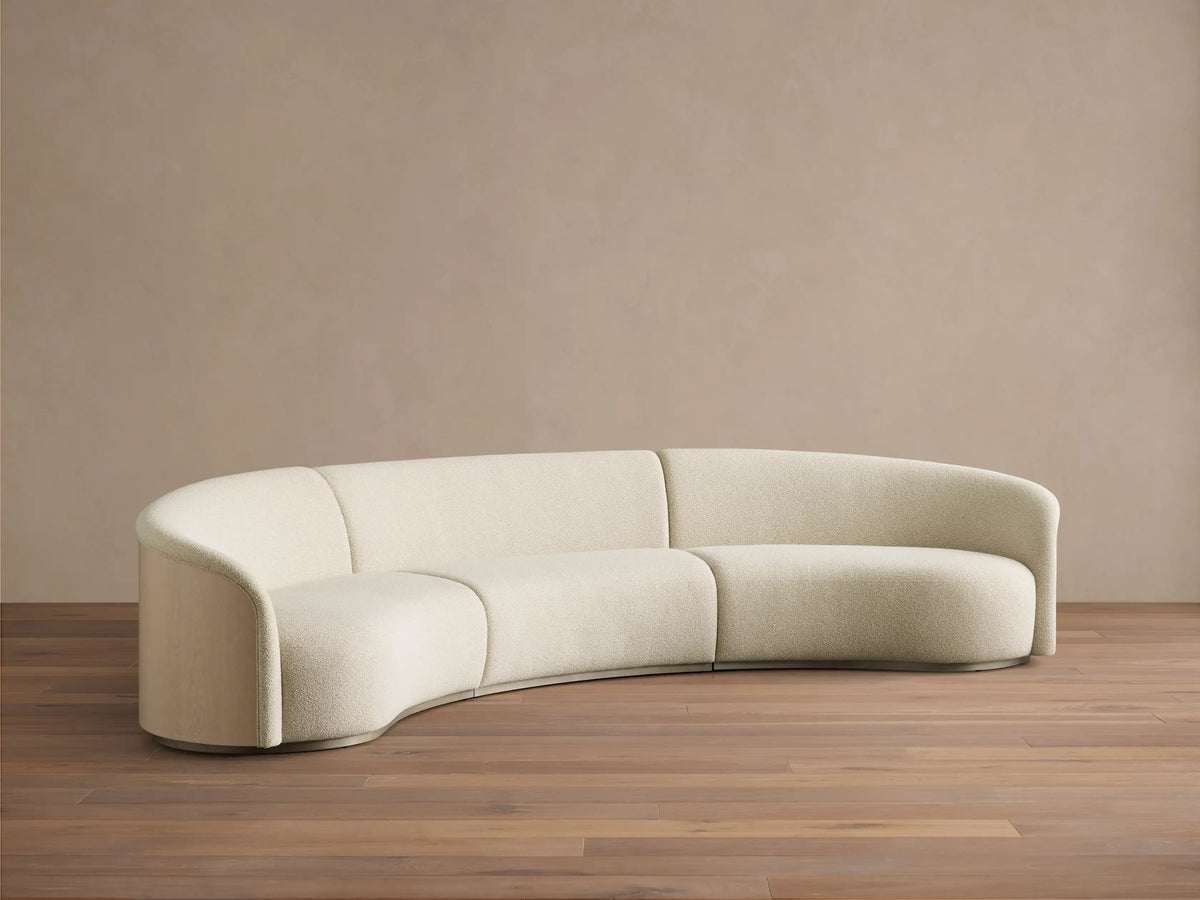 Front view of the curved cream Caden Three-Piece Sectional with sand oak veneer paneling and brushed nickel plinth.