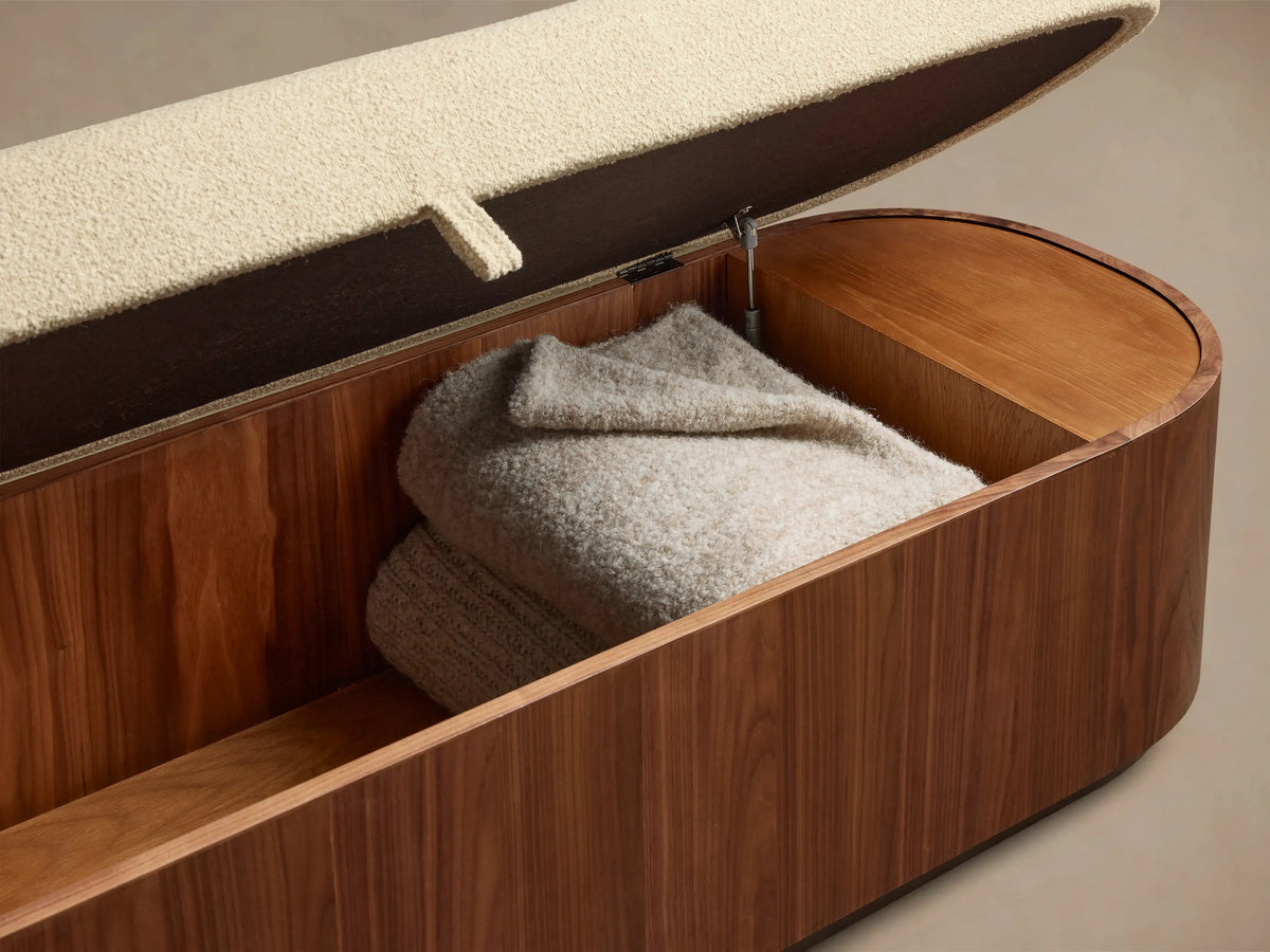 Front view of the Caden Storage Bench, featuring a dark walnut veneer base, an antique bronze plinth, and a light cream upholstered top.
