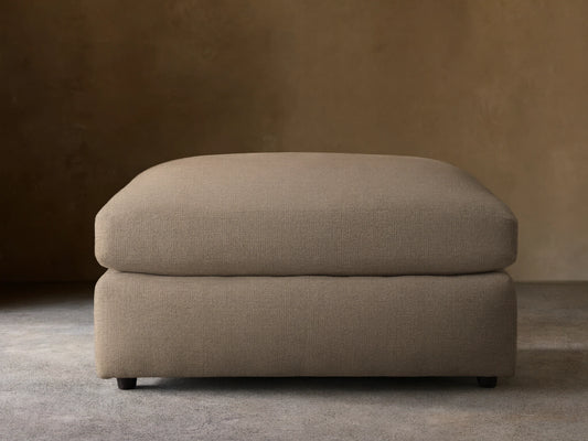 A Kipton Ottoman in soft, light brown textured fabric, featuring a plush, rounded top cushion and a sturdy rectangular base with small dark feet.
