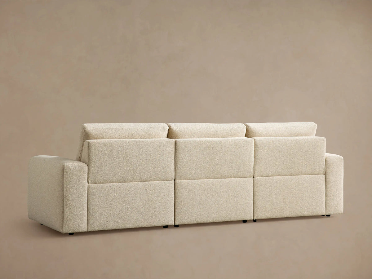 Brennan Three-Piece Motion Sofa in textured off-white fabric. Right seat reclined with footrest extended, against a light brown wall.