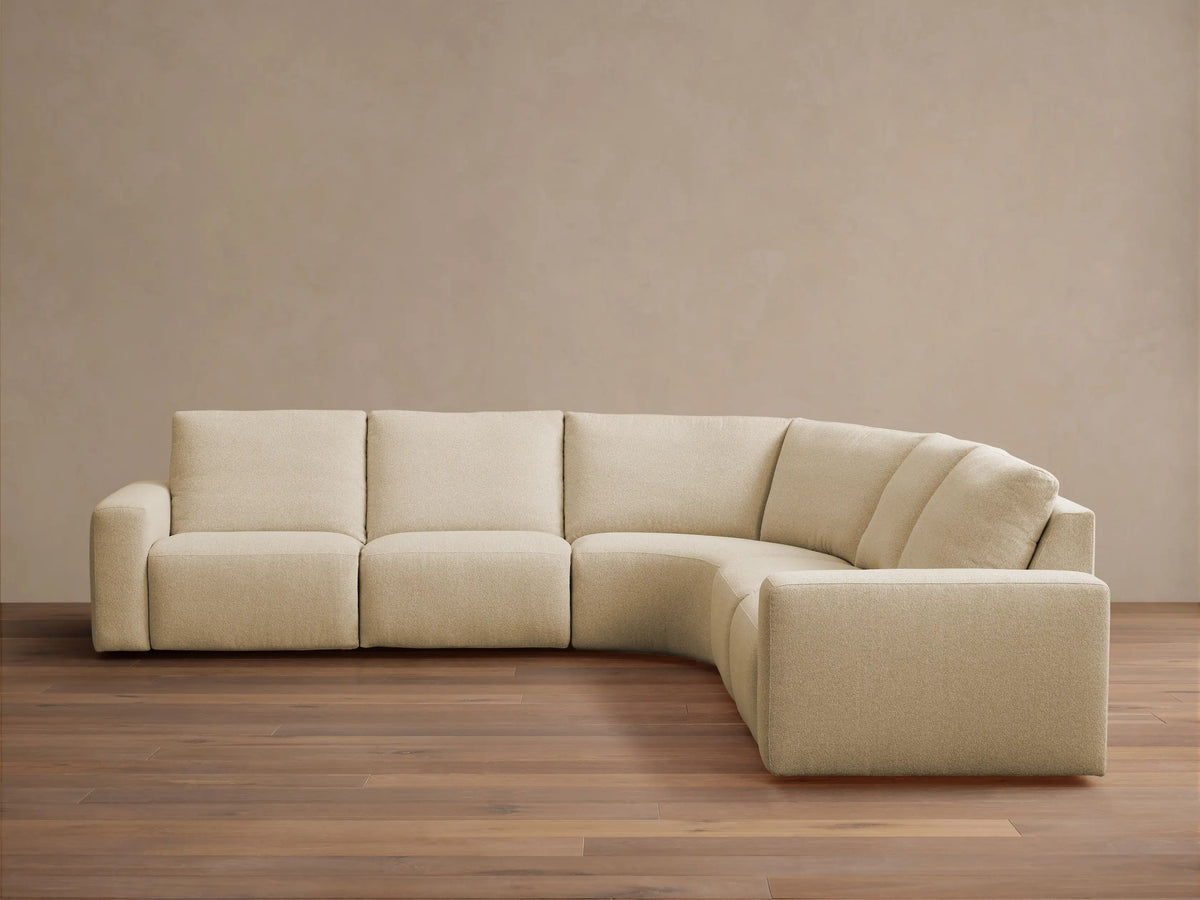 Brennan Five-Piece Motion Corner Sectional: light beige L-shaped sofa with curved corner against a neutral wall, wood floor.