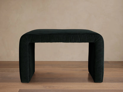 Ryan Ottoman in Banks Midnight, dark upholstered finish, rectangular shape, curved corners, sits on a wooden floor, neutral background.
