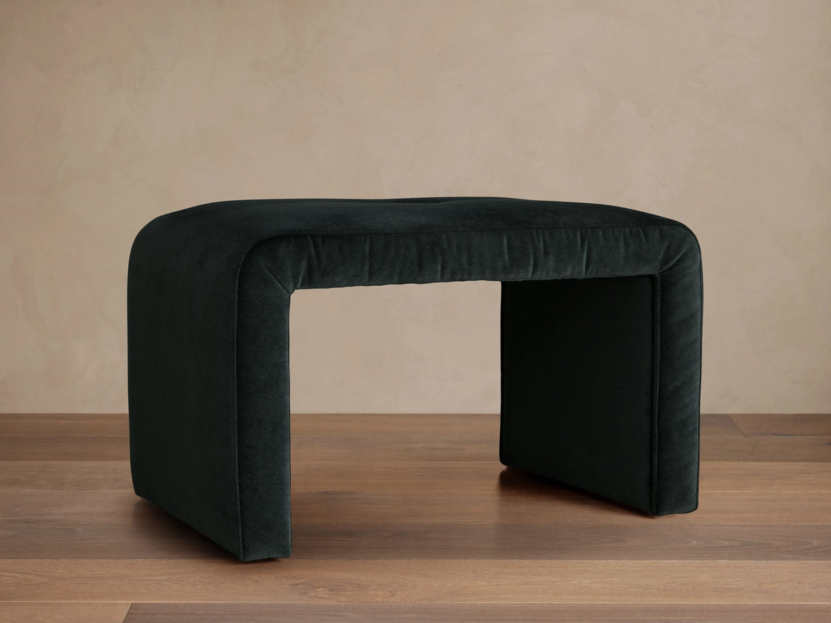 Ryan Ottoman in Banks Midnight, dark upholstered finish, rectangular shape, curved corners, sits on a wooden floor, neutral background.