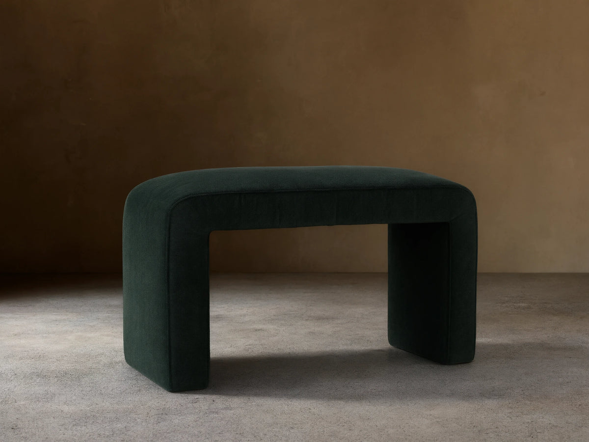 Ryan Ottoman in Banks Midnight, dark upholstered finish, rectangular shape, curved corners, sits on a wooden floor, neutral background.