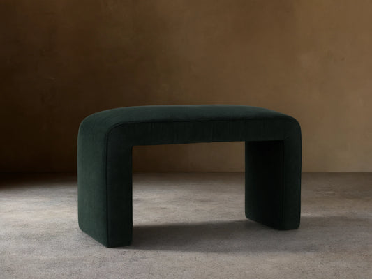 Ryan Ottoman in Banks Midnight, dark upholstered finish, rectangular shape, curved corners, sits on a wooden floor, neutral background.