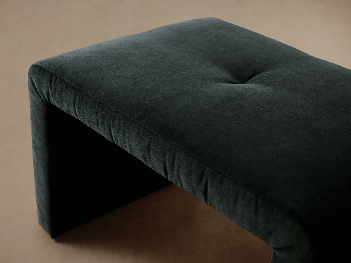 The Ryan Ottoman in Banks Midnight features a dark, rich velvet fabric. It has a sleek, minimalist design with a U-shaped silhouette, sitting on a wooden floor.