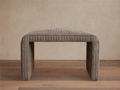Ryan Ottoman featuring a rectangular shape with curved corners. Upholstered in a textured, multicolored fabric, resting on a wooden floor against a neutral wall.