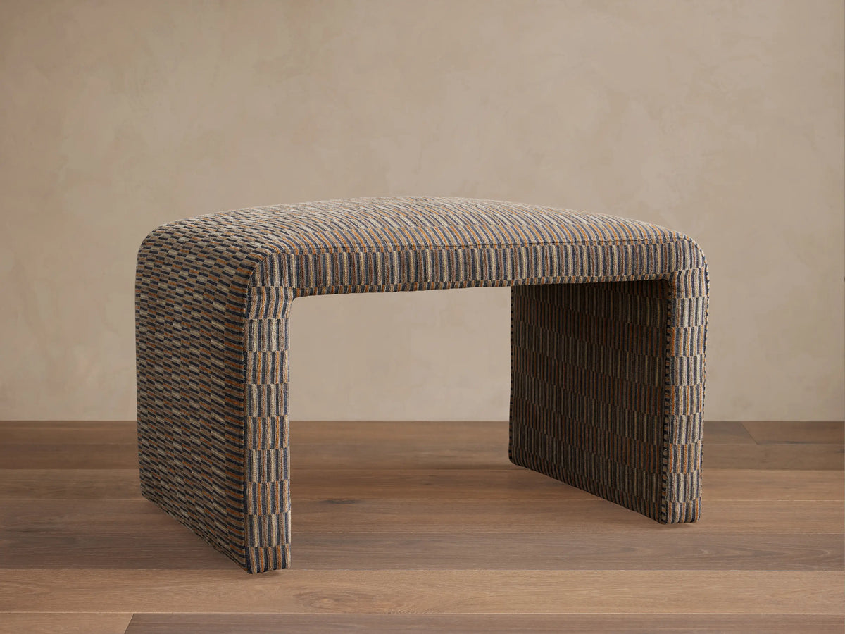 Ryan Ottoman featuring a rectangular shape with curved corners. Upholstered in a textured, multicolored fabric, resting on a wooden floor against a neutral wall.
