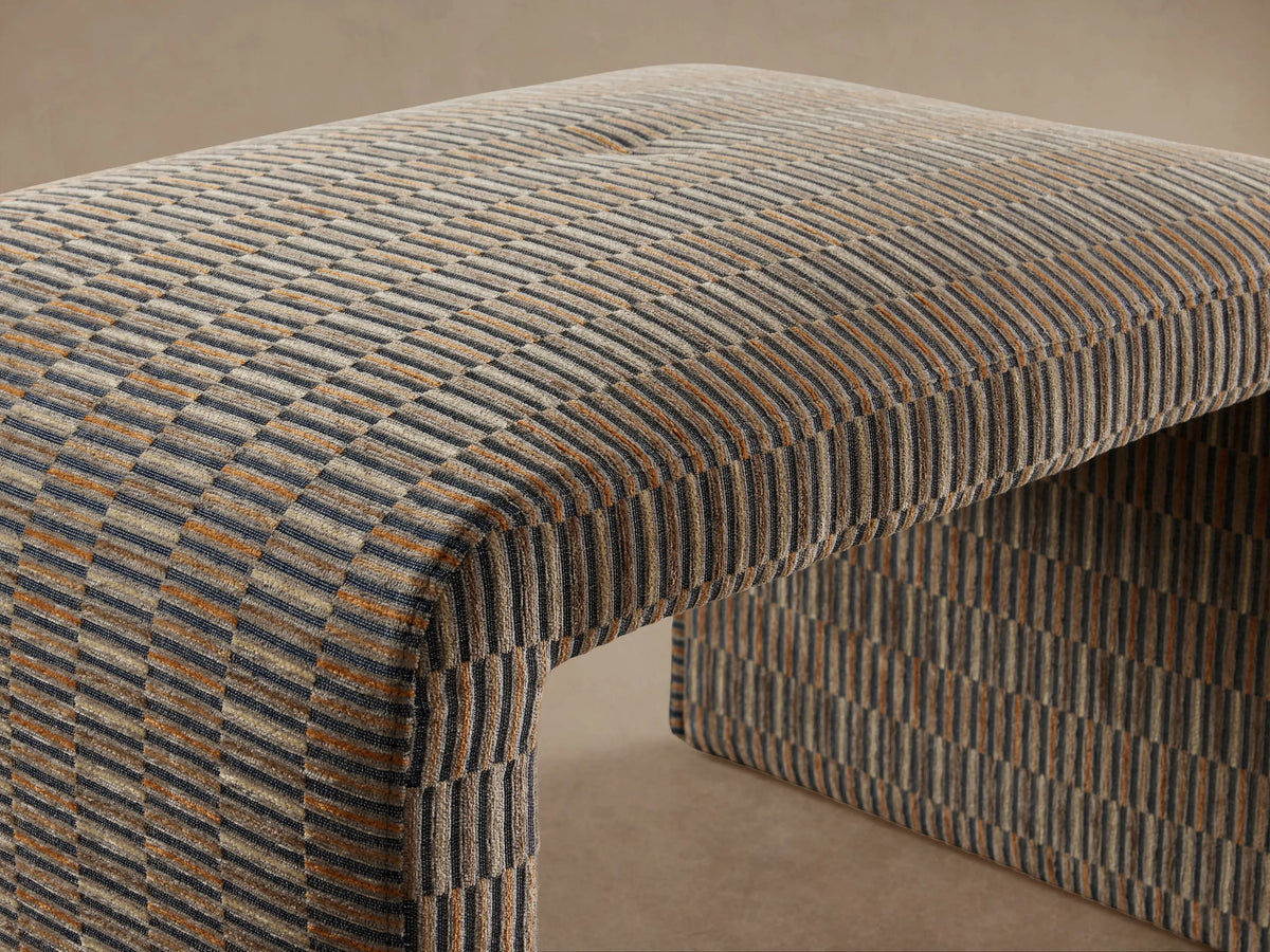 Ryan Ottoman with a woven, textured fabric in a multi-tonal pattern of brown and gray hues, arched top, and straight sides on hardwood floor background.