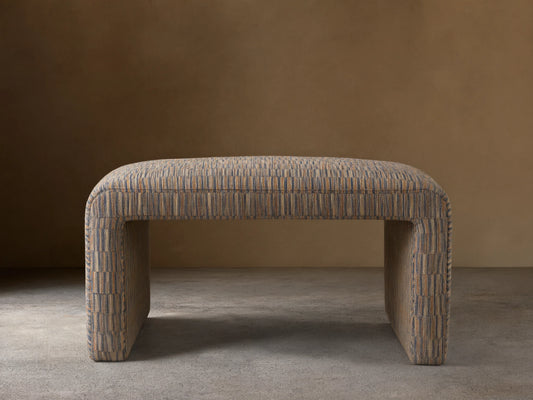 Ryan Ottoman featuring a rectangular shape with curved corners. Upholstered in a textured, multicolored fabric, resting on a wooden floor against a neutral wall.