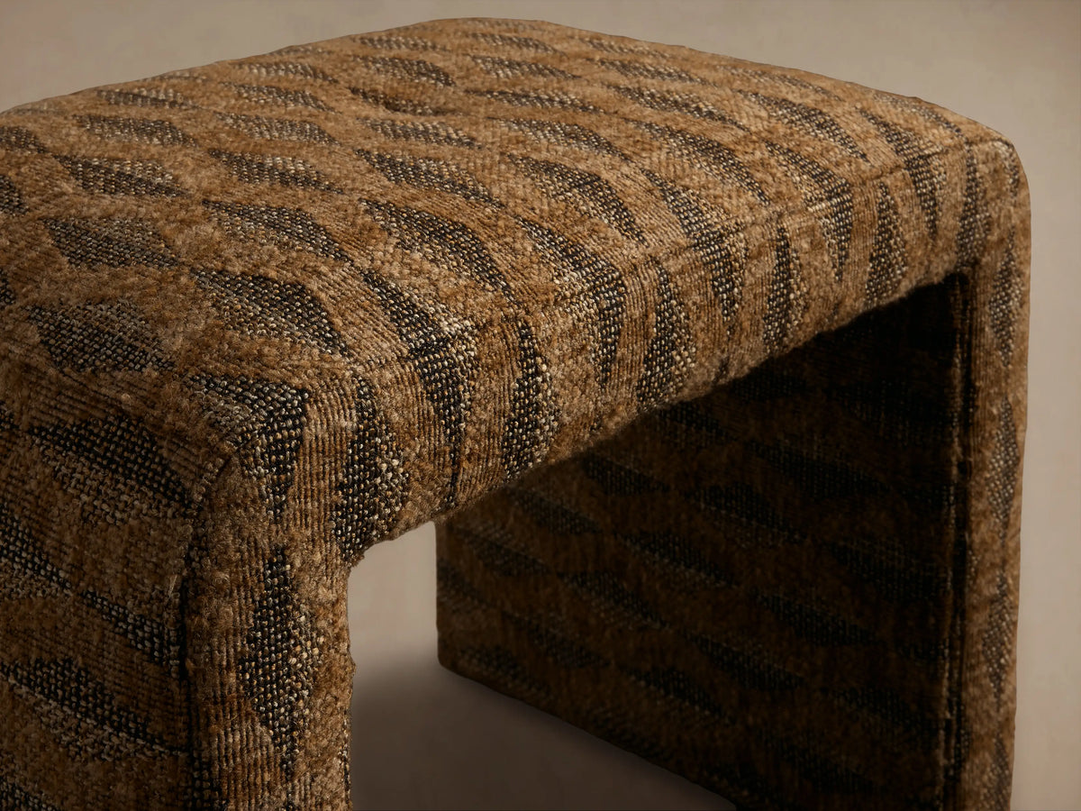 Front view of the Ryan Petite Ottoman, upholstered in a brown and black woven striped fabric with rounded U-shape.