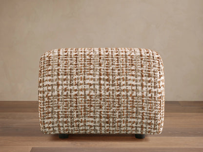 Rectangular ottoman with textured brown and white checkered fabric, placed on a wooden floor, against a beige wall.