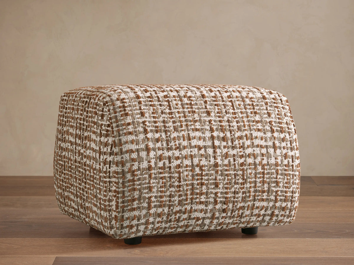 Front view of a Fresno Ottoman featuring a beige, brown, and gray woven checkered pattern, on a patterned rug.