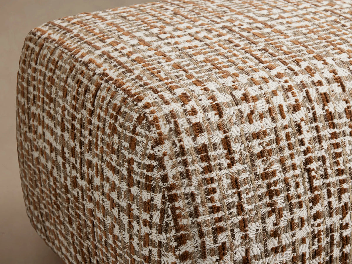 A textured, woven ottoman in shades of brown and cream on a wooden floor, against a neutral beige background.