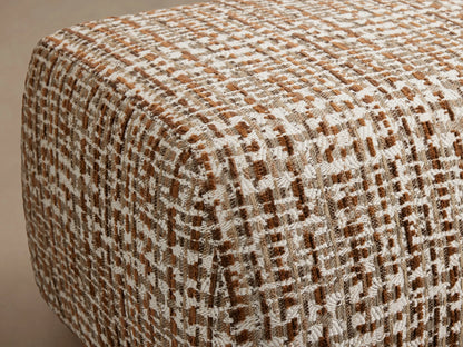 A textured, woven ottoman in shades of brown and cream on a wooden floor, against a neutral beige background.