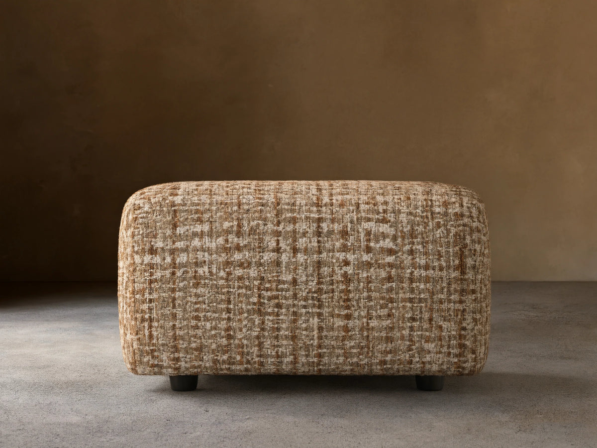 Rectangular ottoman with textured brown and white checkered fabric, placed on a wooden floor, against a beige wall.
