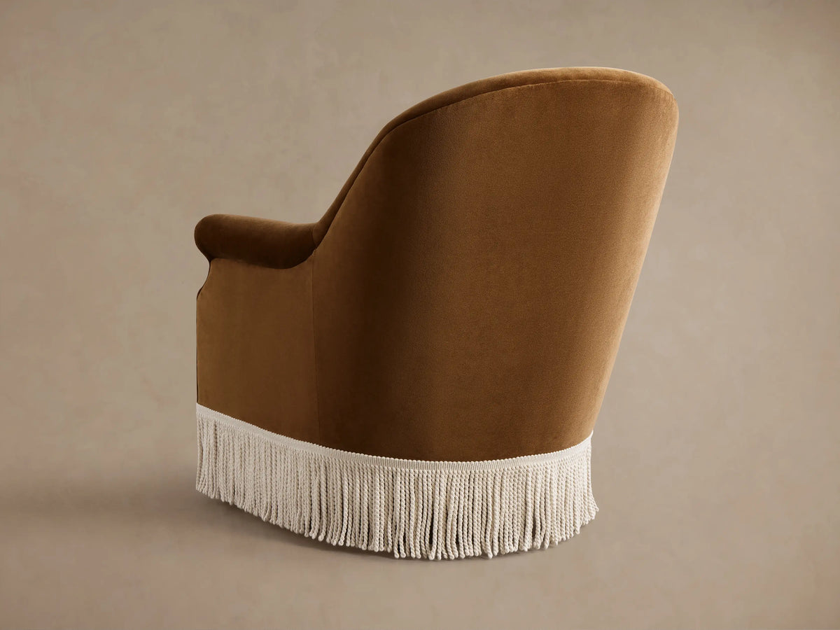 Close-up of the brown Augustus Chair with Fringe, showing its curved arms, seat, and cream fringe along the base.