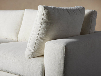 Beale Five Piece Sectional, L-shaped, light cream sofa with plush cushions & ottoman, on wooden floor against a beige wall.