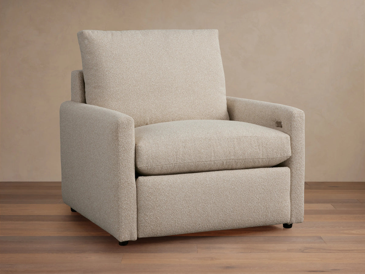 Kipton Motion Recliner in light beige fabric with slim rectangular arms, plush seat, and back cushion on a wooden floor.