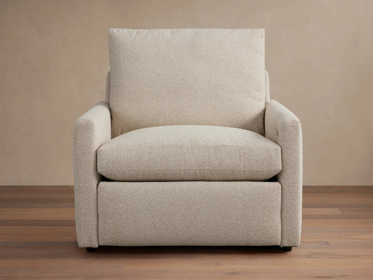 Kipton Motion Recliner in light beige fabric with slim rectangular arms, plush seat, and back cushion on a wooden floor.